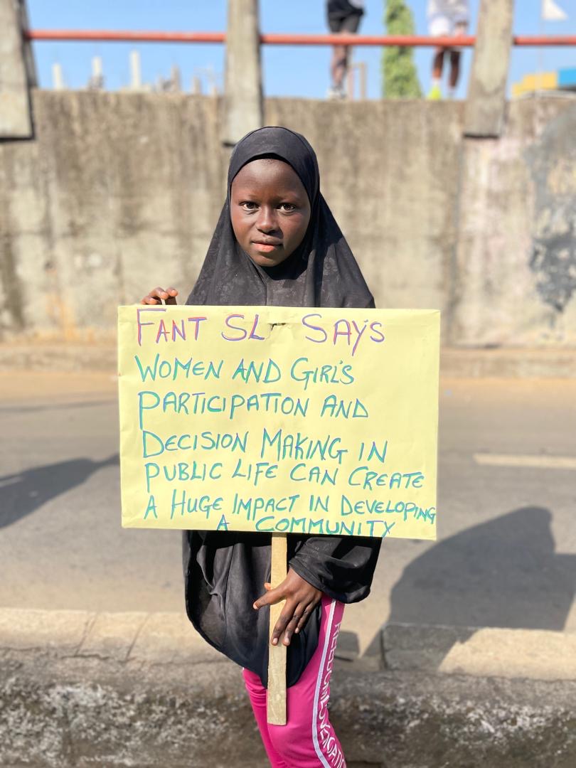 En ung pige fra Sierra Leone står med et skilt, hvorpå der er skrevet: "FANT SL says: Women and girls participation and decision making in public life can create a huge impact in developing a community."